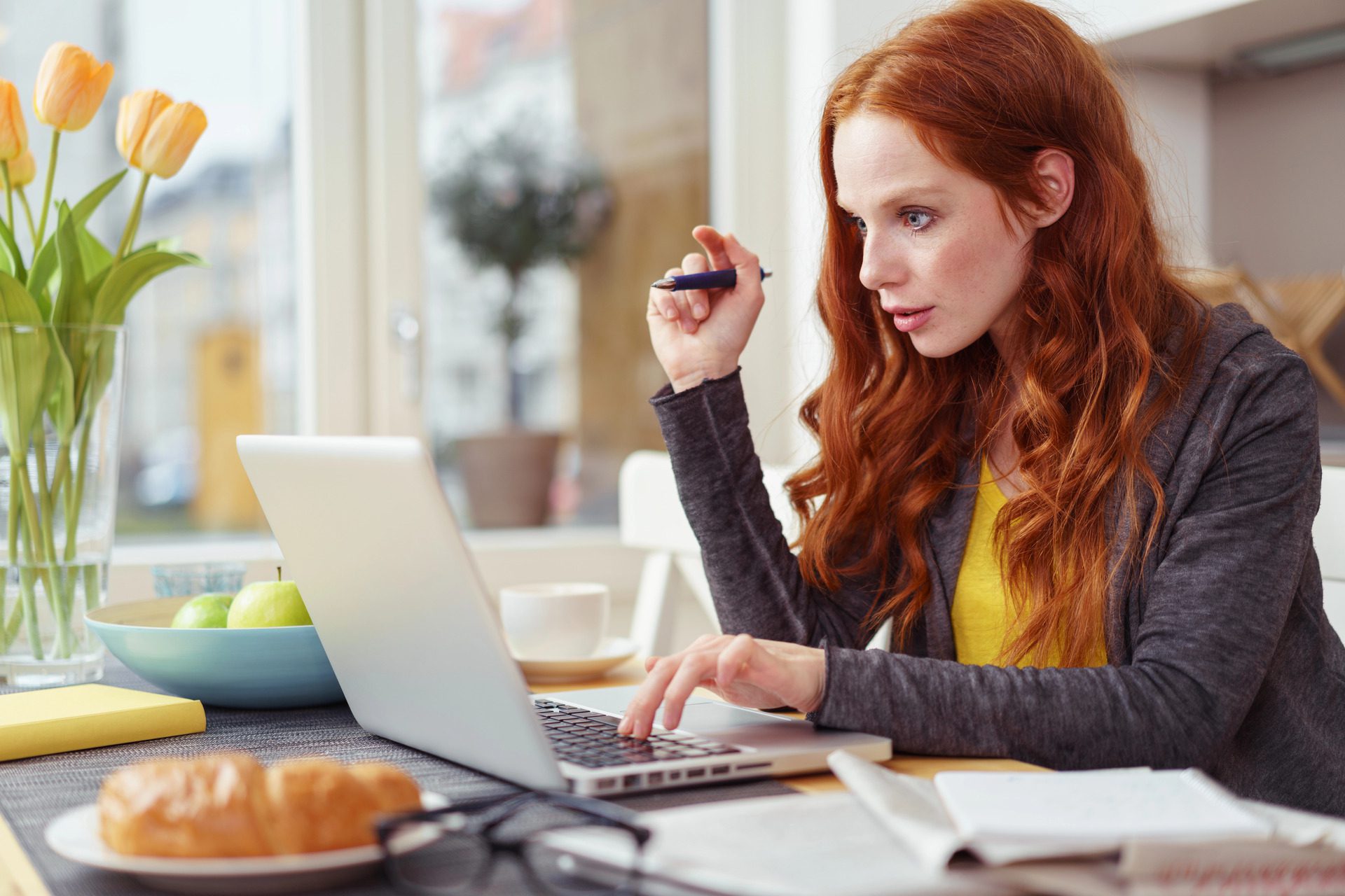 A woman with long red hair works intently on a laptop at a table with a notebook and pen, surrounded by fruit, flowers, and a croissant.